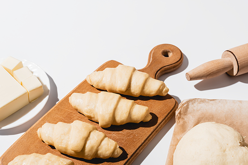 fresh croissants on wooden cutting board near raw dough, rolling pin, butter on white background