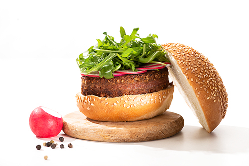delicious vegan burger with radish and arugula on wooden board near black pepper on white background