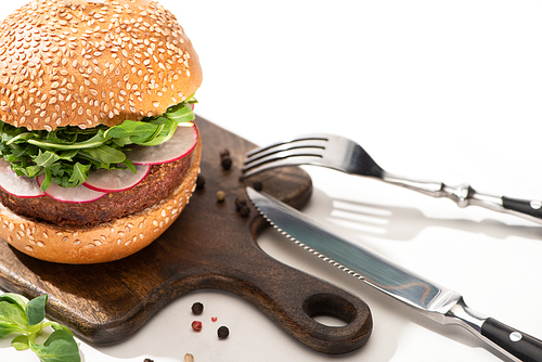 selective focus of delicious vegan burger with radish and arugula on wooden board with black pepper near fork and knife on white background