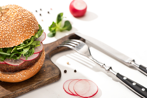selective focus of delicious vegan burger with radish and arugula on wooden board with black pepper near fork and knife on white background