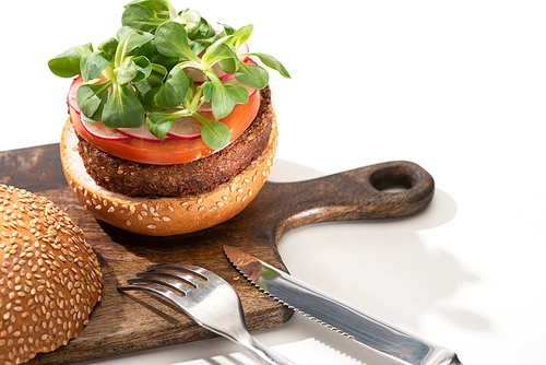 delicious vegan burger with radish, tomato and microgreens on wooden boar near fork and knife on white background