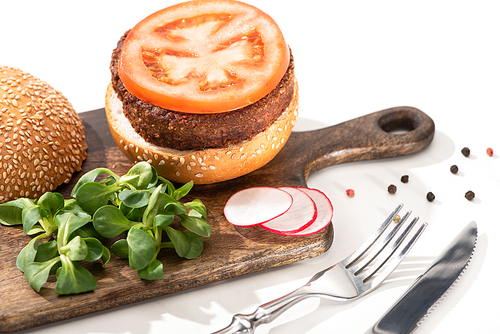 selective focus of delicious vegan burger with radish, tomato and microgreens on wooden boar with black pepper near fork and knife on white background