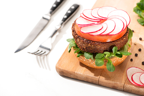 selective focus of delicious vegan burger with radish, tomato and microgreens on wooden boar with black pepper near fork and knife on white background