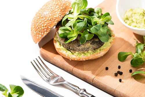 delicious green vegan burger with microgreens and mashed avocado on wooden boar with black pepper near fork and knife on white background