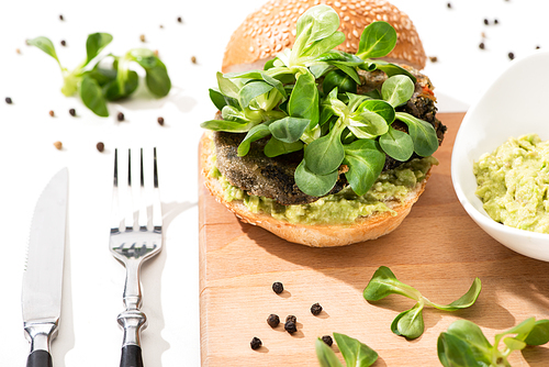 delicious green vegan burger with microgreens and mashed avocado on wooden boar with black pepper near fork and knife on white background