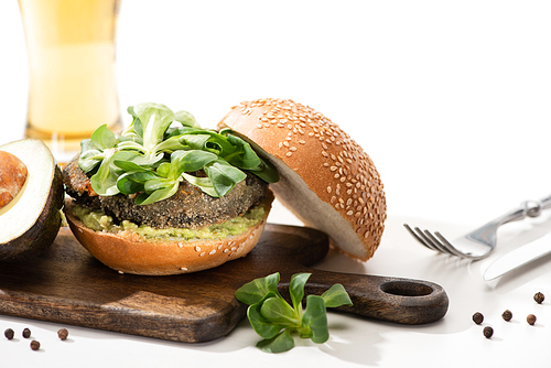 selective focus of delicious green vegan burger with microgreens, avocado, black pepper on wooden cutting board near fork and oil on white background