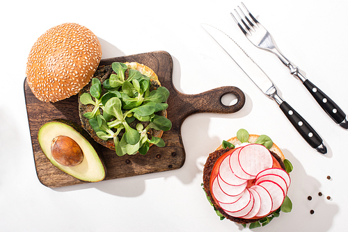 top view of vegan burgers with microgreens, avocado, radish on wooden cutting board on white background with cutlery
