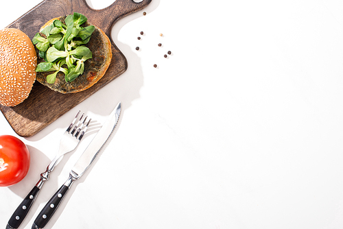 top view of vegan burger with microgreens on wooden board near tomato and cutlery on white background