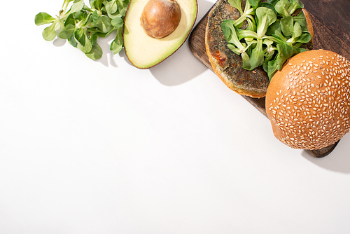 top view of vegan burger with microgreens and avocado on wooden cutting board on white background