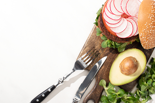 top view of delicious vegan burger with radish, avocado and greens on wooden cutting board with cutlery on white background