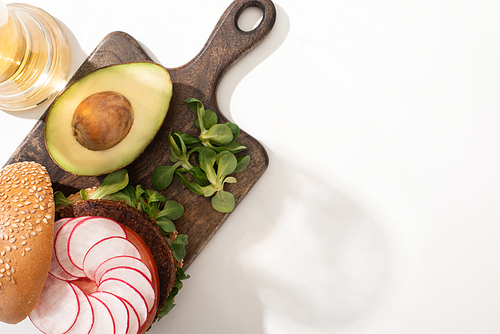 top view of delicious vegan burger with radish, avocado and greens on wooden cutting board on white background