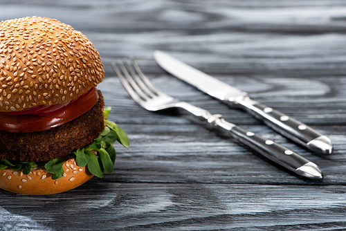 selective focus of tasty vegan burger with vegetables served on wooden table with fork and knife