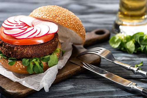 selective focus of tasty vegan burger with radish and tomato served on cutting board with cutlery on wooden table