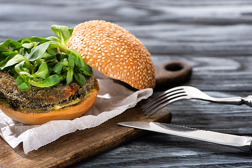 tasty vegan burger with microgreens served on cutting board with cutlery on wooden table
