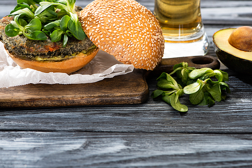 tasty vegan burger with microgreens served on cutting board near avocado and beer on wooden table
