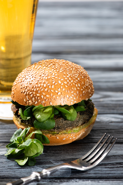 selective focus of tasty vegan burger with microgreens served on wooden table with glass of beer and fork