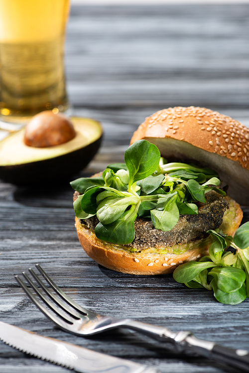 selective focus of tasty vegan burger with microgreens served on wooden table with avocado half, cutlery and beer