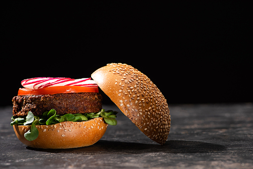 tasty vegan burger with microgreens, radish and tomato served on textured surface isolated on black