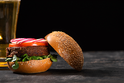 selective focus of tasty vegan burger with microgreens, radish and tomato served on textured surface with beer isolated on black