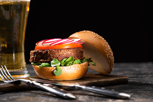 selective focus of tasty vegan burger with microgreens, radish and tomato served with beer and cutlery on textured surface isolated on black