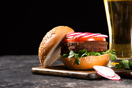 selective focus of tasty vegan burger with s served on wooden cutting board near beer isolated on black