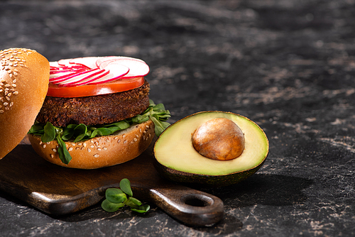 tasty vegan burger with vegetables served with avocado half on wooden cutting board on textured surface