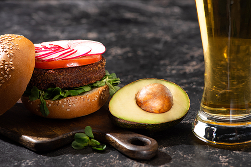 tasty vegan burger with vegetables served with avocado half and beer on wooden cutting board on textured surface