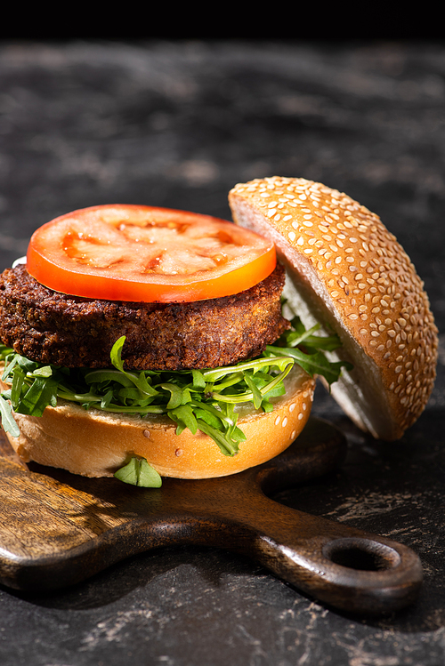 tasty vegan burger with tomato and greens served on wooden cutting board on textured surface
