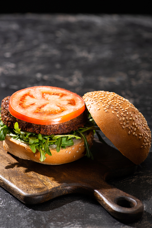 selective focus of tasty vegan burger with tomato and greens served on wooden board on textured surface