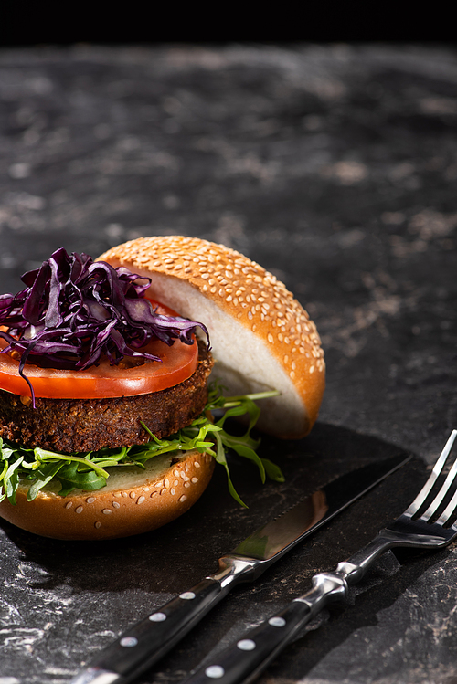 tasty vegan burger with tomato, red cabbage and greens served on textured surface with cutlery