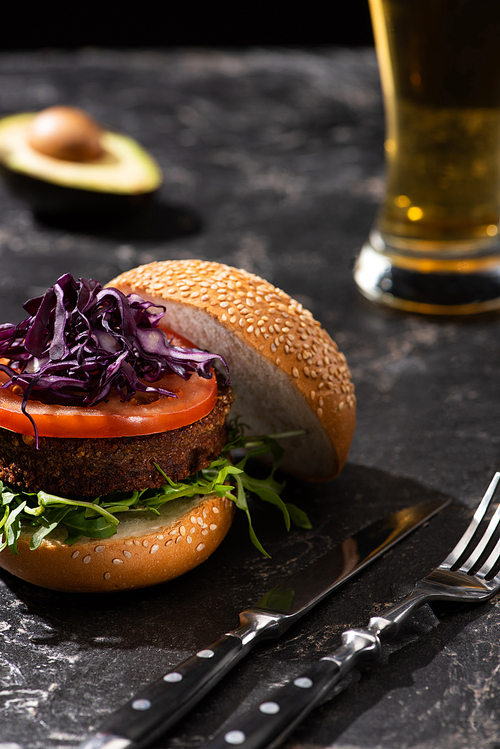 selective focus of tasty vegan burger with tomato, red cabbage and greens served on textured surface with cutlery, beer and avocado