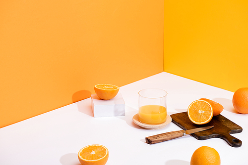 fresh orange juice in glass near ripe oranges, wooden cutting board with knife on white surface on orange background