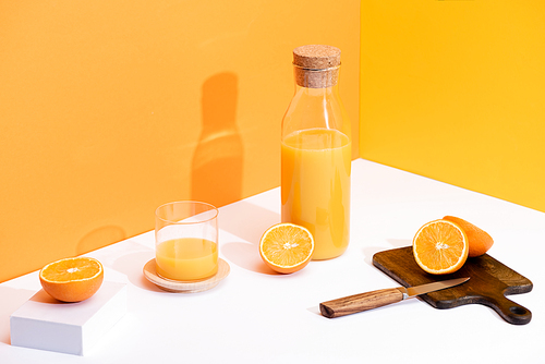 fresh orange juice in glass and bottle near ripe oranges, wooden cutting board with knife on white surface on orange background