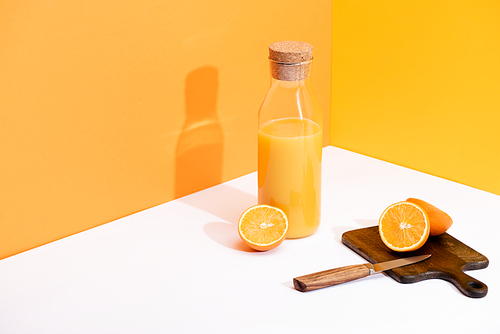 fresh orange juice in glass bottle near ripe oranges, wooden cutting board with knife on white surface on orange background