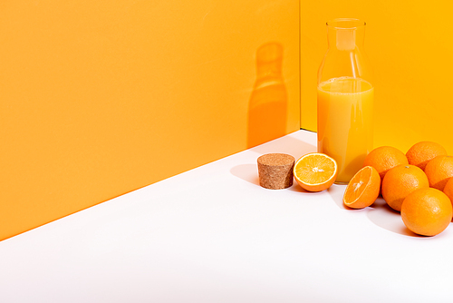 fresh orange juice in glass bottle near ripe oranges and cork on white surface on orange background