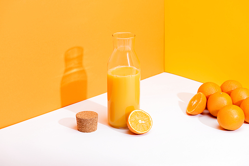 fresh orange juice in glass bottle near ripe oranges and cork on white surface on orange background