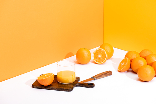 fresh orange juice in glass on wooden cutting board with knife near ripe oranges on white surface on orange background