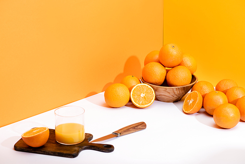fresh orange juice in glass on wooden cutting board with knife near ripe oranges in bowl on white surface on orange background