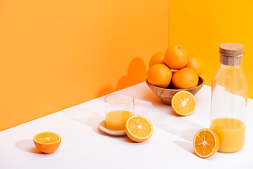 fresh orange juice in glass and bottle near ripe oranges in bowl on white surface on orange background