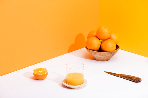 fresh orange juice in glass near ripe oranges in bowl and knife on white surface on orange background