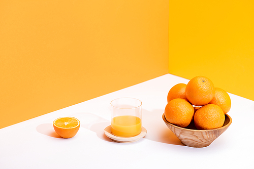 fresh orange juice in glass near ripe oranges in bowl on white surface on orange background