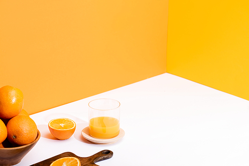 fresh orange juice in glass near ripe oranges in bowl and wooden cutting board on white surface on orange background