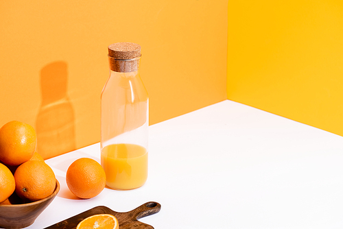 fresh orange juice in glass bottle near ripe oranges in bowl and cutting board on white surface on orange background