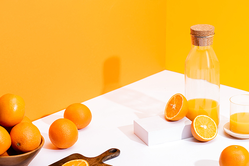 fresh orange juice in glass and bottle near ripe oranges in bowl and cutting board on white surface on orange background