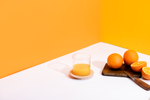 fresh orange juice in glass near ripe oranges on cutting board with knife on white surface on orange background