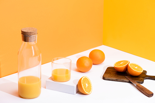 fresh orange juice in glass and bottle near ripe oranges on cutting board with knife on white surface on orange background