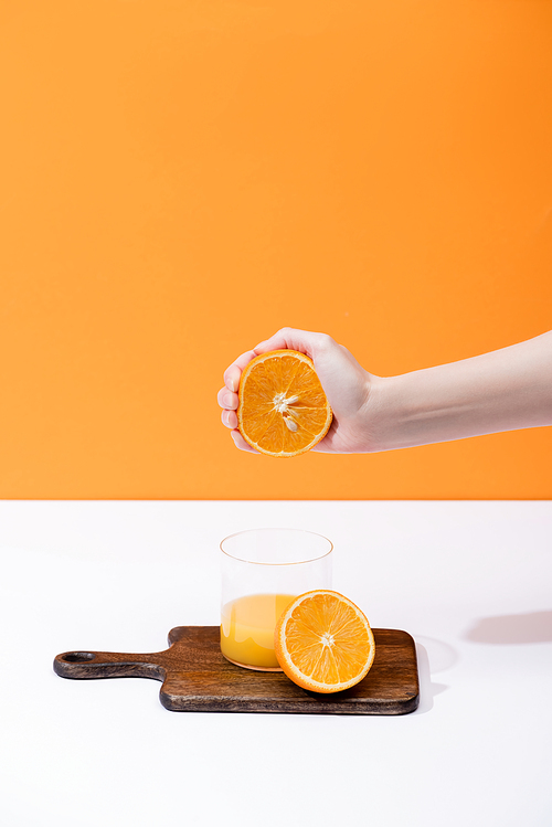 cropped view of woman squeezing fresh orange juice in glass on wooden cutting board on white surface isolated on orange