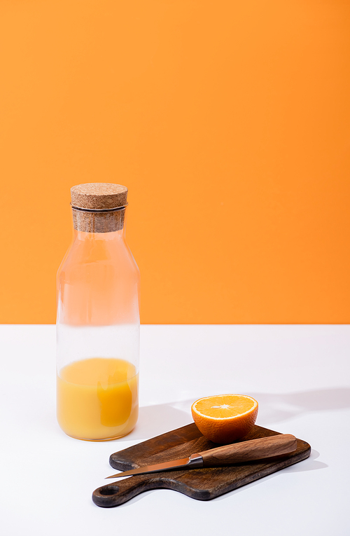 fresh orange juice in glass bottle near cut fruit on wooden cutting board with knife on white surface isolated on orange