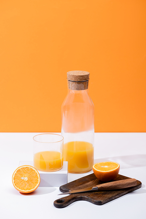 fresh orange juice in glass and bottle near cut fruit on wooden cutting board with knife on white surface isolated on orange