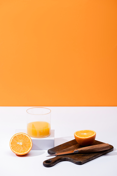 fresh orange juice in glass near cut fruit on wooden cutting board with knife on white surface isolated on orange